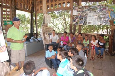 Hombre dirige una charla a un grupo de niños en una choza de madera.