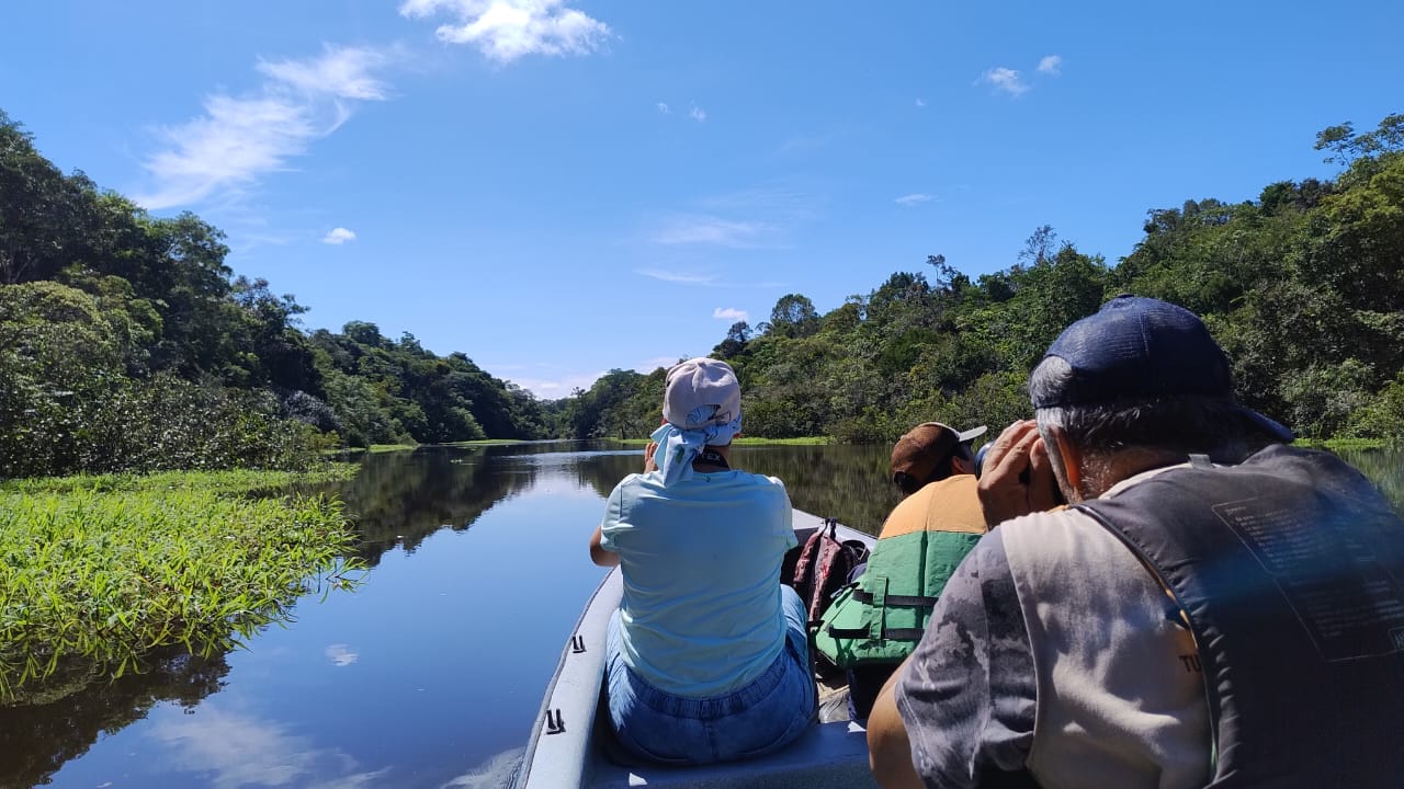 Tres personas navegan un río en canoa durante el día, árboles y cielo azul en segundo plano.