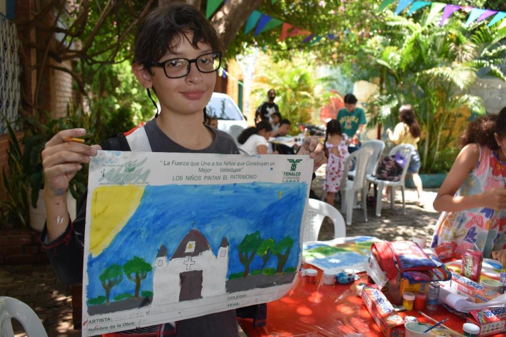 Niño exhibe una acuarela con otros niños pintando en segundo plano.